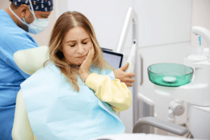 editor-1780645530013-17710605 Woman in a dental chair clutches her jaw in discomfort while a dentist in scrubs and a mask works in the background.