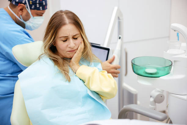 Woman in a dental chair clutches her jaw in discomfort while a dentist in scrubs and a mask works in the background.